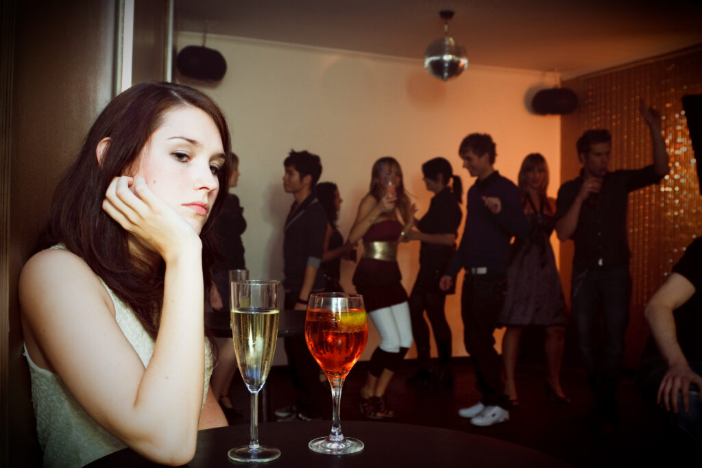 young sad woman is sitting alone in a nightclub while a crowd of people is dancing in the background. two drinks are standing on the table in front of her.