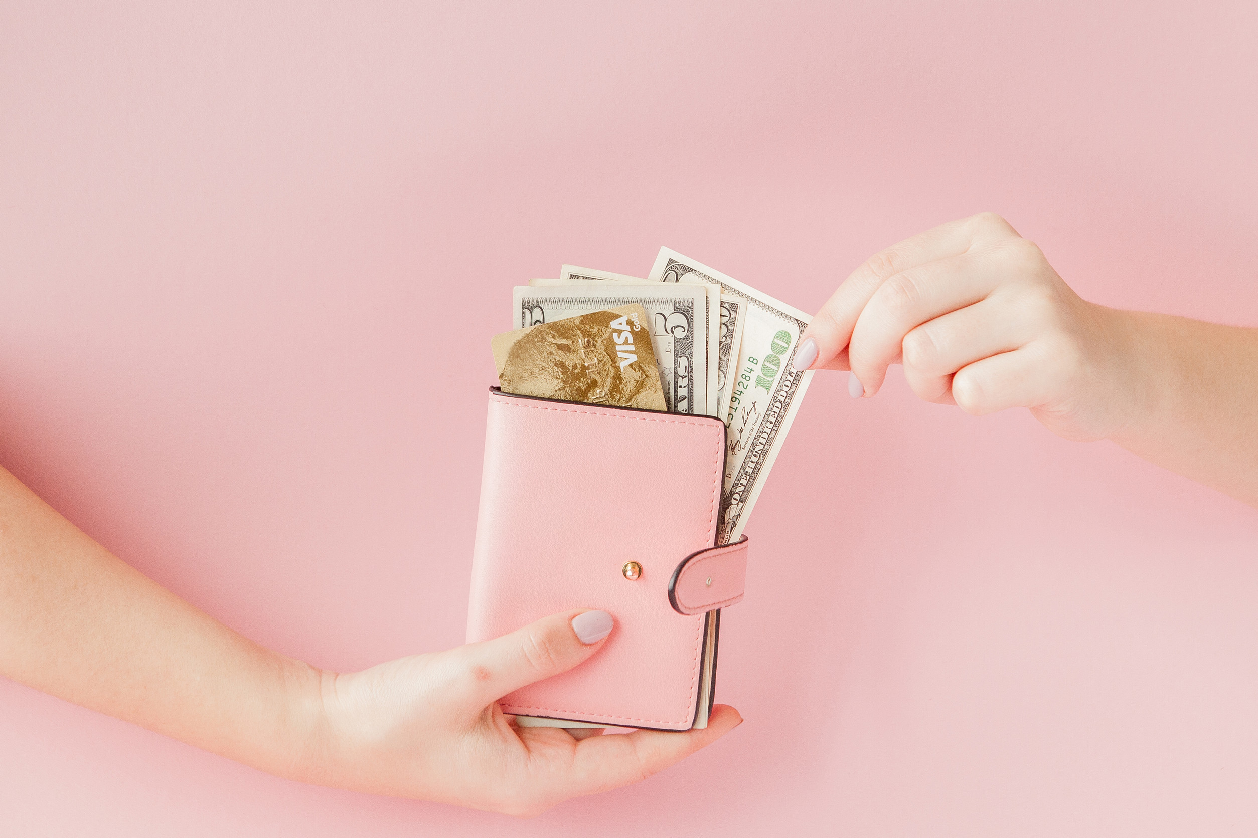 Dollars and pink wallet with credit card in woman's hands on pink background.