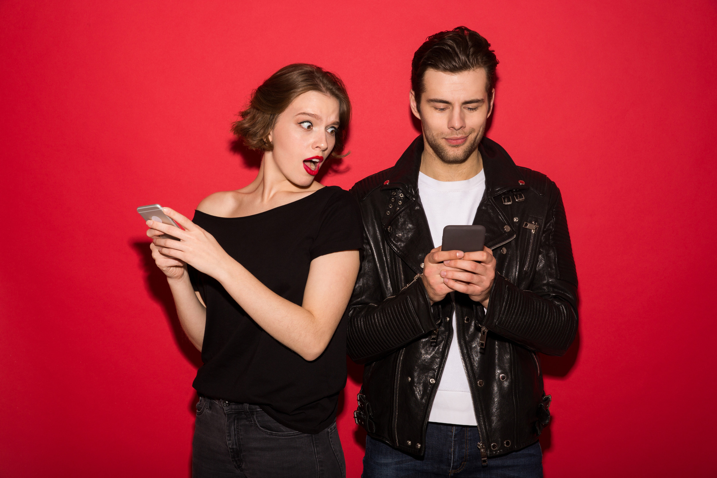 Smiling male punk using smartphone while woman peeps at him over red background